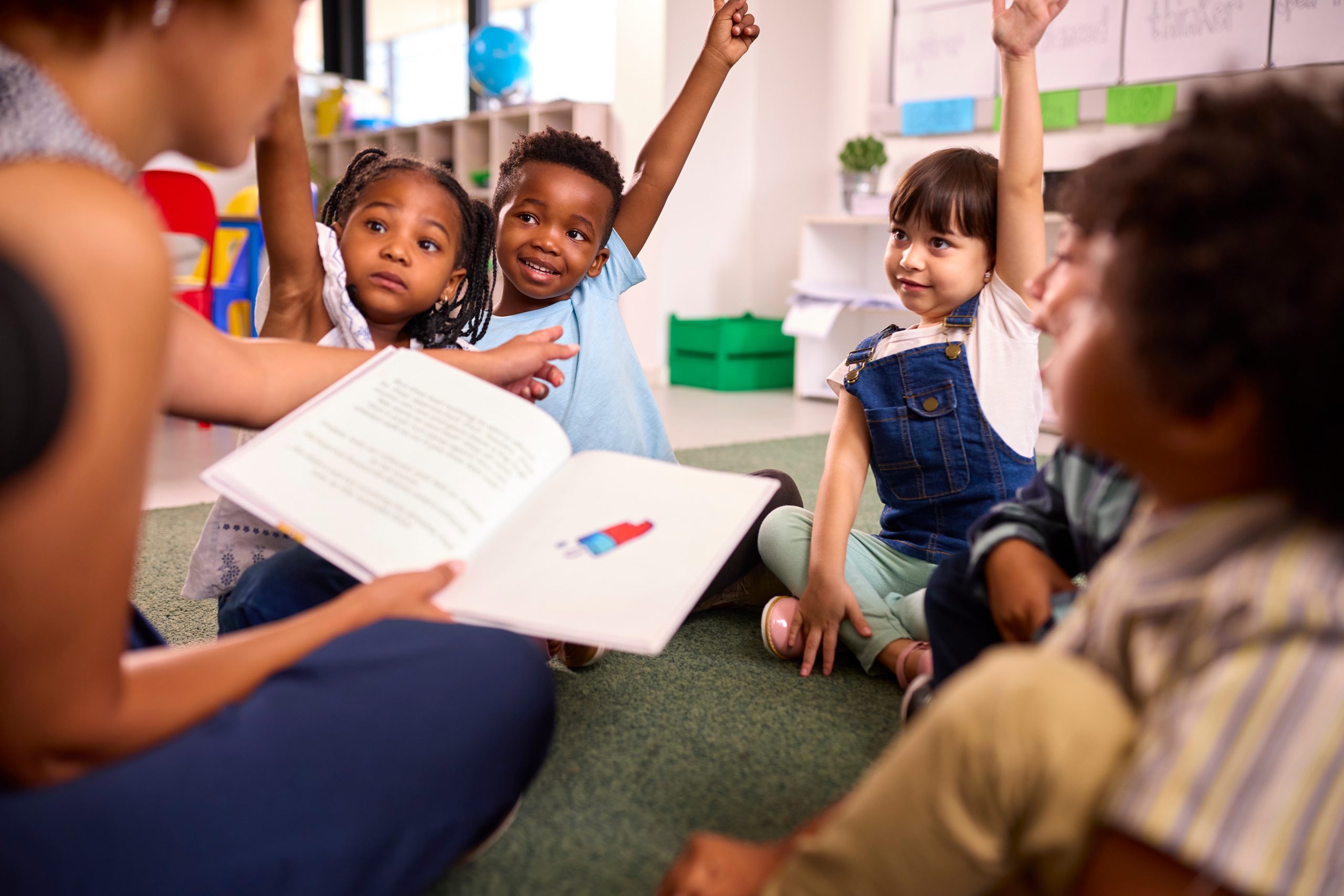 A teacher reads to elementary school students sitting in a circle on the floor with their hands raised to answer a question.